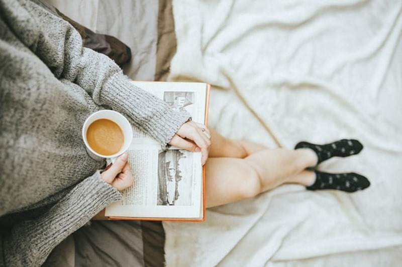Apprendre à lire plus Jeune femme assise sur son lit avec un livre sur les jambes et un café à la main.