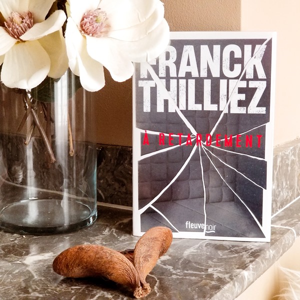 Franck Thilliez - A retardement Photo du livre "A retardement" de Franck Thilliez posé sur le rebord d'une cheminée avec des fleurs blanches dans un vase