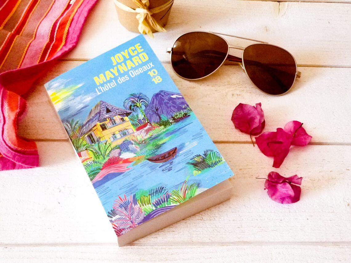 Photo du livre "L'hôtel des oiseaux" de Joyce Maynard posé sur une table en bois blanc avec lunettes de soleil et fleurs de bougainvilliers rose