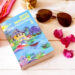 Photo du livre "L'hôtel des oiseaux" de Joyce Maynard posé sur une table en bois blanc avec lunettes de soleil et fleurs de bougainvilliers rose